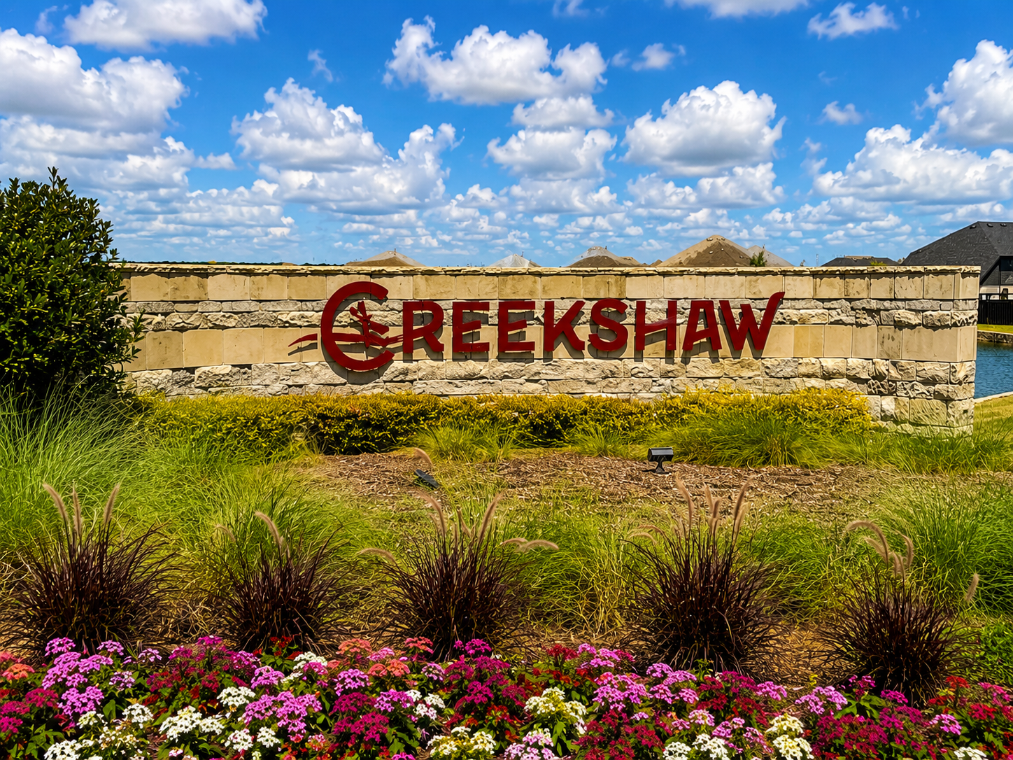 Stone sign reading Creekshaw in red letters on a beige stone wall, with a landscaped garden bed in front and a blue, cloud-filled sky behind.
