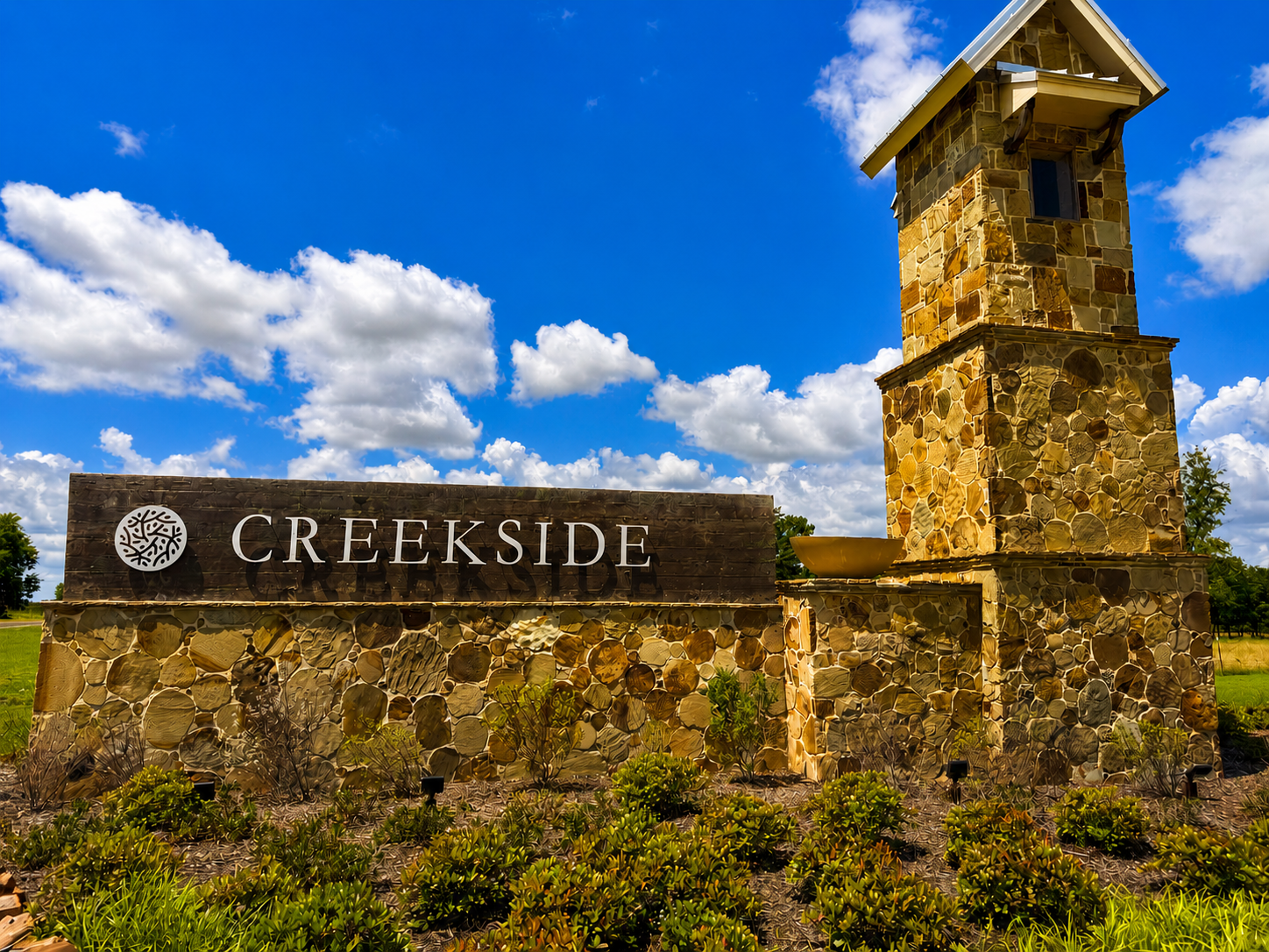 Stone Creekside entrance sign mounted on a stone wall with a tall stone tower, under a blue sky with white clouds.