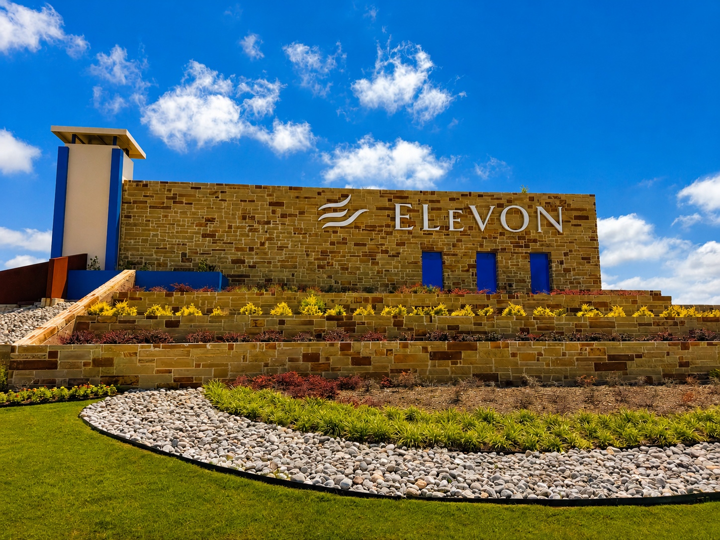 Stone building with 'ELEVON' sign on a tan brick wall, blue windows, and layered landscaping with rocks and plants under a bright blue sky