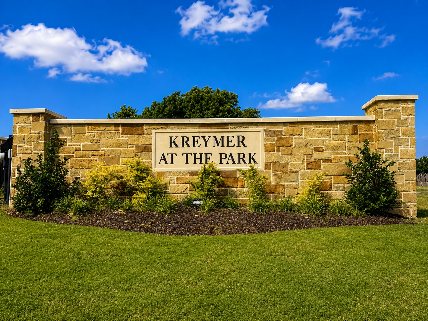 Stone entrance sign reading 'KREYMER AT THE PARK' with landscaped bushes and a bright blue sky backdrop