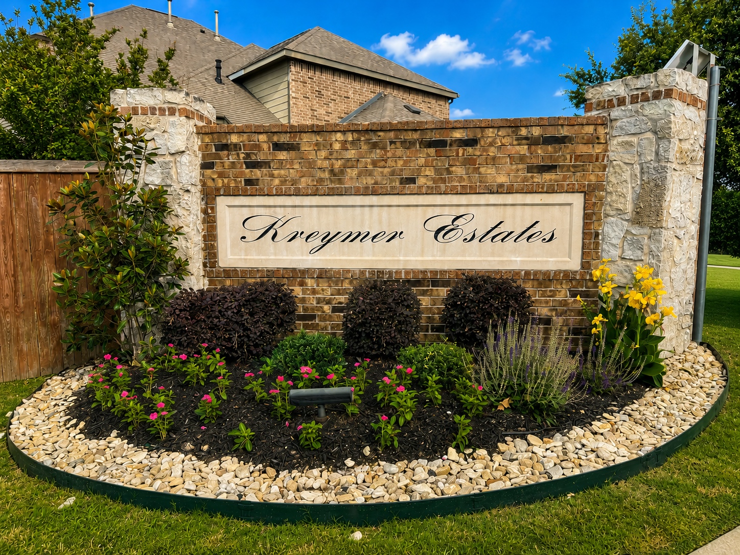 Brick and stone entrance sign reading 'Kreymer Estates' with a landscaped bed of yellow lilies, pink flowers, and purple shrubs under a blue sky.