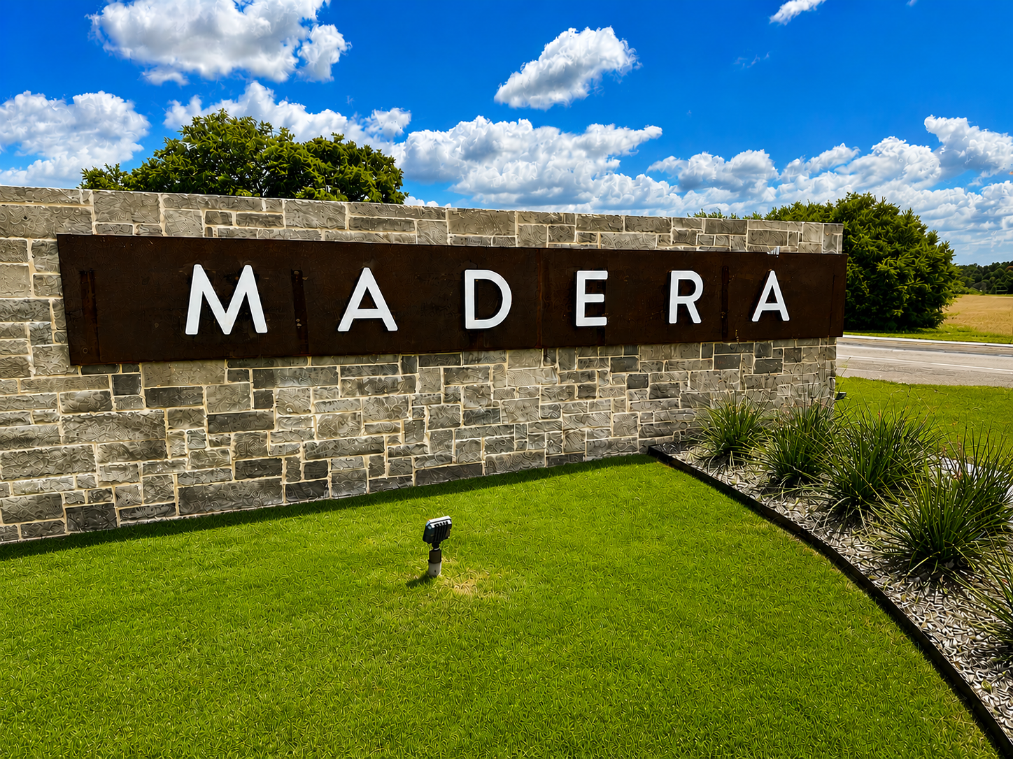 Stone entry sign reading MADERA mounted on a textured stone wall with green lawn and a blue sky with clouds behind it.