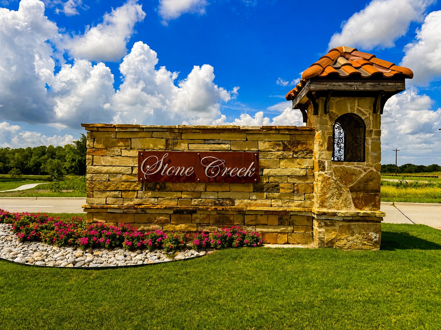 Stone Creek entrance sign on a stone wall with a small tower and red-tiled roof, set against a blue sky with clouds.