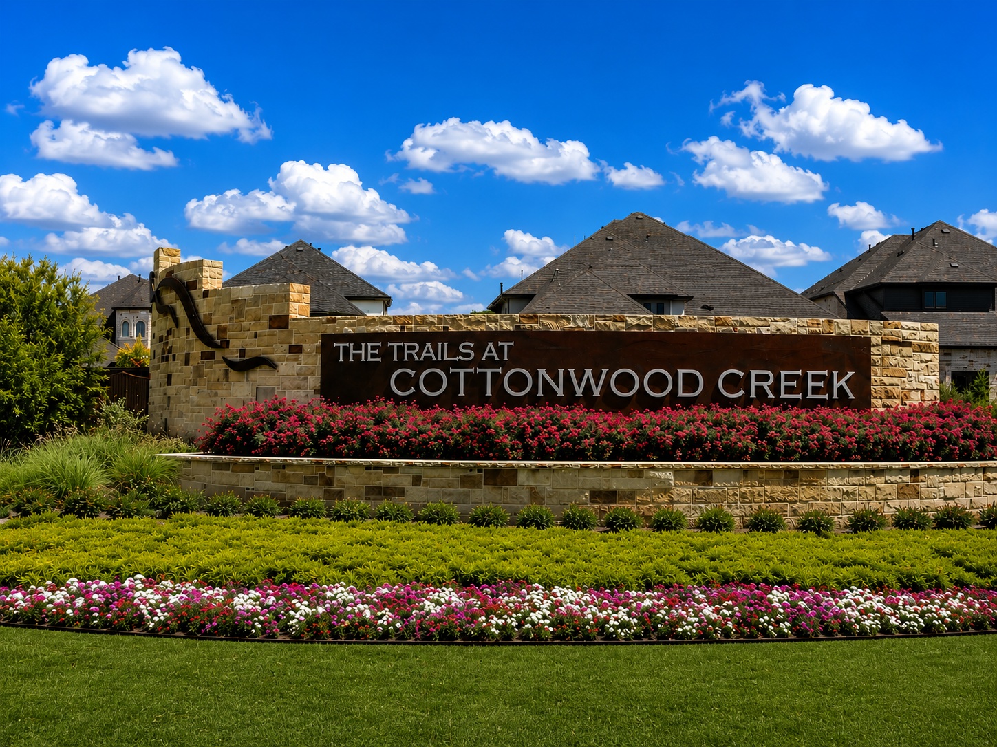 Stone entrance sign reading 'The Trails at Cottonwood Creek' with red flowering beds and green shrubs under a blue sky.