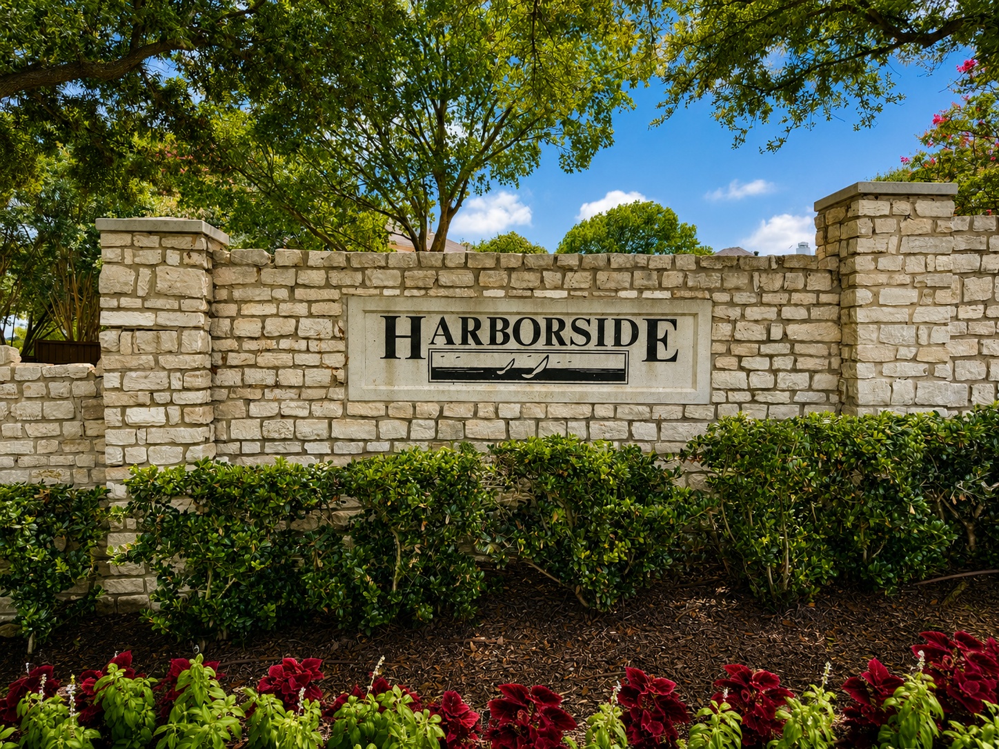 Stone wall entrance with a sign reading HARBORSIDE, framed by green shrubs and red flowers under a bright blue sky.